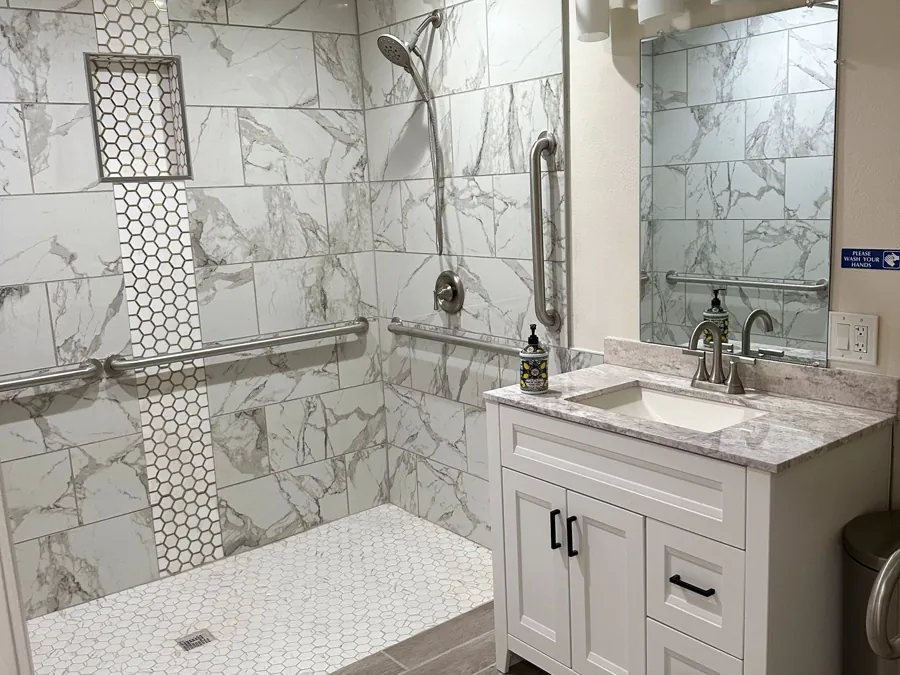 A step-free tiled shower with grab bars and a handheld shower head, beside a vanity